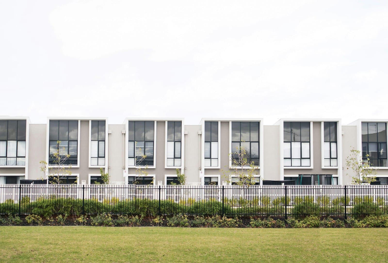 A row of narrow, modern rendered townhouses with black-framed windows behind a steel picket fence.
